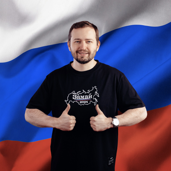 Улыбающийся Андрей Замай на фоне флага Российской Федерации, показывающй жест «ОК»